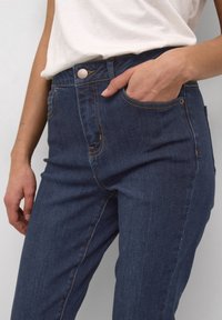 Persona vistiendo jeans azul oscuro de cintura alta, con la mano parcialmente dentro del bolsillo delantero y una camiseta blanca metida por dentro.