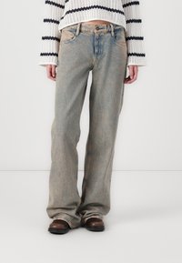 Jeans de mezclilla azul claro con un corte relajado, textura deslavada y un sutil efecto de desgaste. Combinados con calzado marrón y un suéter de punto a rayas.