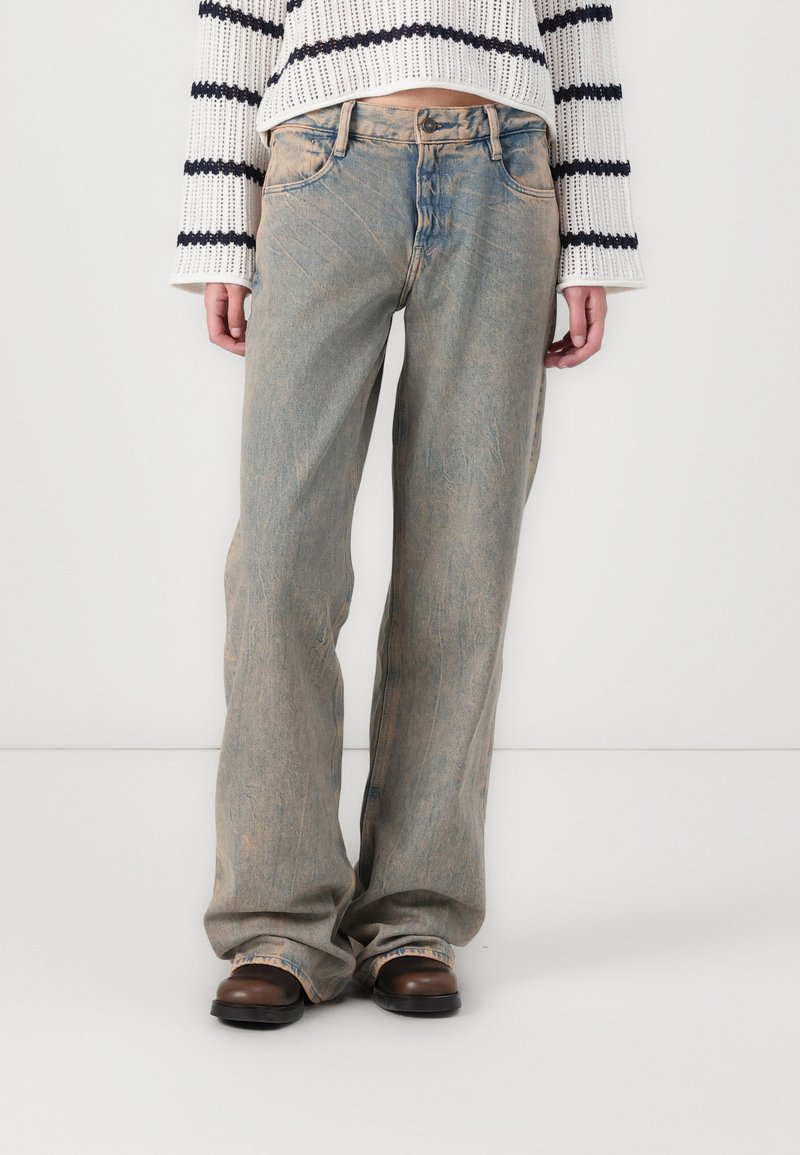 Jeans de mezclilla azul claro con un corte relajado, textura deslavada y un sutil efecto de desgaste. Combinados con calzado marrón y un suéter de punto a rayas.