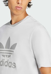 Bílé tričko s krátkým rukávem vyrobené z bavlny, s velkým šedým logem Adidas na přední straně, kulatým výstřihem a žebrovaným límcem.