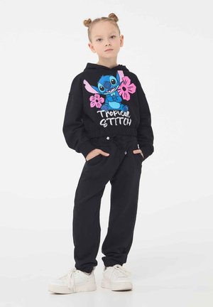 Felpa nera con grafica "Tropical Stitch" e accenti floreali, abbinata a pantaloni jogger neri. Polsi elasticizzati e tasche. Sneakers bianche.