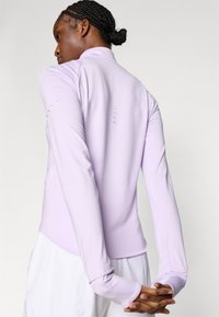 Μωβ μωβ αθλητικό τοπ με μακριά μανίκια, ελαφρώς καμπυλωτό τελείωμα, επίπεδες ραφές και διακριτικές λεπτομέρειες αερισμού στο πίσω μέρος. Ελαφρύ, ελαστικό υλικό.