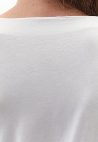 Bílý, lehký látkový top s hladkou texturou a širokým výstřihem, odhalující záda s jednoduchým, minimalistickým designem.