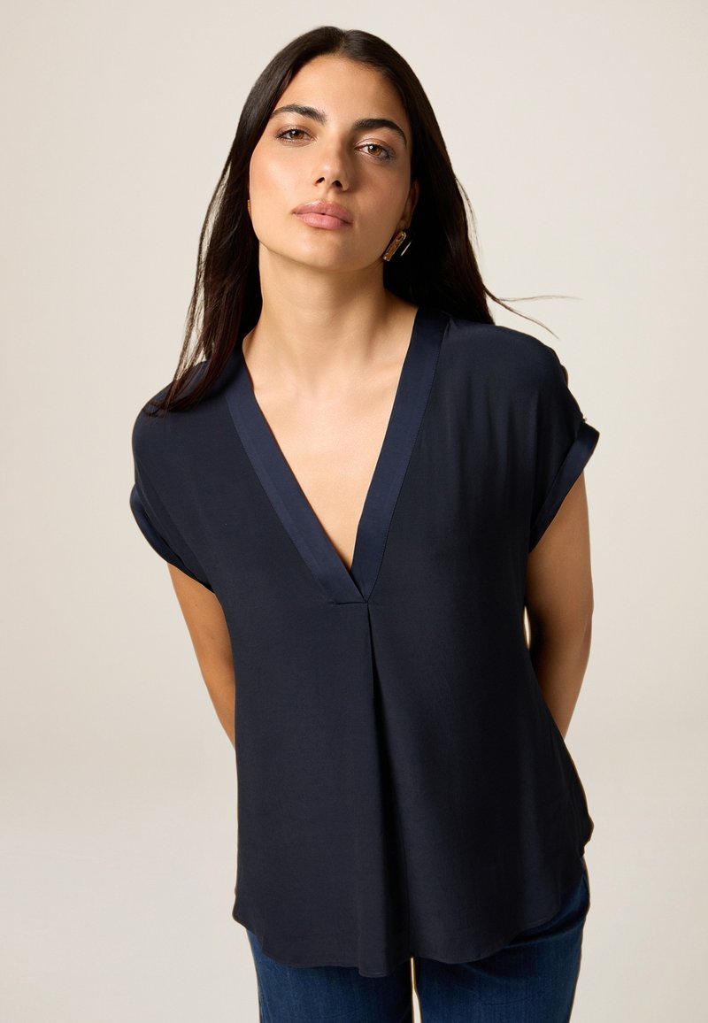 Blouse à manches courtes bleu marine avec un décolleté en V profond, en tissu doux et à coupe décontractée, présentant un éclat subtil et un détail de couture au centre.