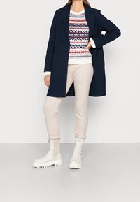 Manteau bleu marine à coupe structurée, pull à motifs rouges, bleus et crème, joggeurs beiges, et bottines blanches à semelle épaisse.