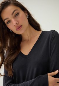 Crni pleteni pulover s V-izrezom, dugim rukavima, glatke teksture i rebrastim detaljima na izrezu. Model je prikazan na neutralnoj pozadini.