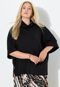 Top nero oversize con collo a scialle e maniche corte, realizzato in tessuto liscio, abbinato a una gonna con fantasia. Braccialetto d'oro visibile al polso.