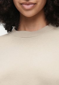 Pulover bej cu guler rotund și margine ondulată, realizat dintr-un material moale, texturat. Vedere de aproape a gulerului și a zonei gâtului.