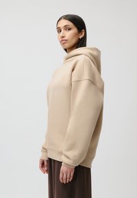 Beige oversized hettegenser laget av mykt stoff med stor hette, dropped shoulders, ribbestrikkede mansjetter, og en avslappet passform. Modellen har den på sammen med et brunt skjørt.