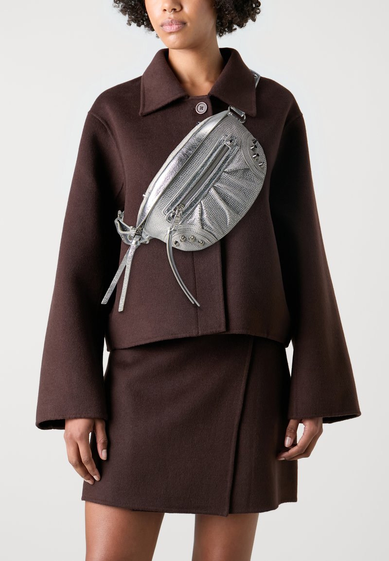 Veste courte en laine marron avec un col, assortie d'une jupe. Sac bandoulière en argent avec zips et clous.