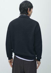 Schwarzer Pullover mit strukturierter Oberfläche, mit rundem Ausschnitt und gerippten Bündchen, über einem weißen Hemd und dunkler Hose getragen.