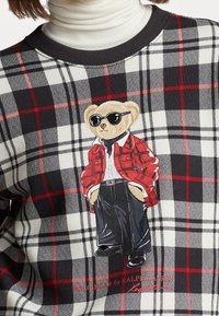 Rot-schwarzer Karo-Pullover mit einem illustrierten Bären in Sonnenbrille, der eine rote Jacke und schwarze Hose trägt. Aus weichem Stoff mit geripptem Kragen.