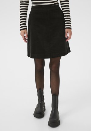 Frau trägt einen schwarzen Cordrock, transparente schwarze Strumpfhosen, schwarze klobige Stiefel und ein langärmeliges schwarz-weiß gestreiftes Oberteil.