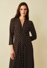 Mallimodellissa on tummanruskea mekko, jossa on valkoisia pilkkuja. Mekossa on syvä kaula-aukko ja pitkät hihat. Hänen takanaan on vaaleanbeige tausta.