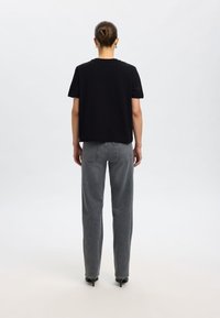 Črna kratka majica s kratkimi rokavi, sproščen kroj, kombinirana z visokim pasom sive kavbojke z ravnimi hlačnicami. Vidni čevlji z visokimi petami. Enostaven ozadje.