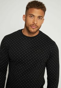 Camisa negra de manga larga con pequeños patrones en blanco y gris de cruces, cuello redondo y textura de tela suave. Detalle del logo sutil en el pecho.