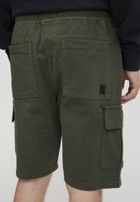 Olivenfargede cargo-shorts med elastisk midjebånd, flere store lommer og et stoffmerke. Laget av bomull med en glattsk.Texture.