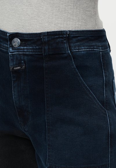 Jean en denim bleu foncé à la taille avec bouton visible et étiquette de marque porté avec un haut côtelé gris clair rentré.