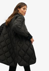 Vrouw in een lange zwarte gewatteerde jas met een golfpatroon, die over haar schouder kijkt tegen een effen witte achtergrond.