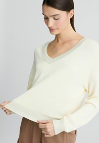 Maglione lavorato a maglia color crema con scollatura a V, orlo e polsini a coste, caratterizzato da un logo discreto sul bordo inferiore. Texture morbida e vestibilità comoda.