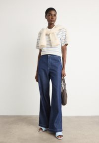 Blaue Jeans mit weitem Bein, gestreiftes Kurzarmoberteil mit gelben und blauen Linien, flauschiger cremefarbener Pullover um den Hals gebunden, blaue Sandalen.