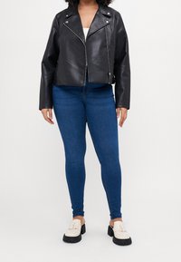Chaqueta de motocicleta de cuero negro con herrajes plateados, diseño ajustado, mangas largas y un largo recortado, combinada con jeans skinny azules y mocasines color crema.