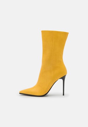 Żółty teksturowany botek na kostkę ze spiczastym noskiem i cienkim czarnym obcasem typu szpilka na białym tle.