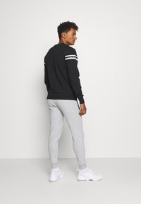 Black sweatshirt with white stripes on shoulders, gray sweatpants, white sneakers; shown from the back, model facing slightly to the side.