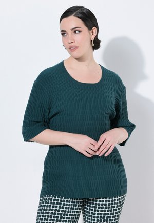 Donna con capelli corti scuri, che indossa un maglione a trama verde scuro e pantaloni a fantasia bianca e nera, che guarda di lato con le mani incrociate.