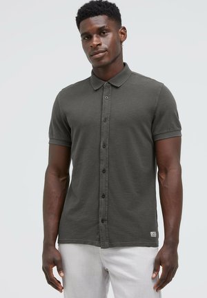 Junger Schwarzer Mann, der ein dunkelgraues kurzärmeliges Hemd und hellgraue Hose trägt, steht vor einem einfarbigen hellen Hintergrund.