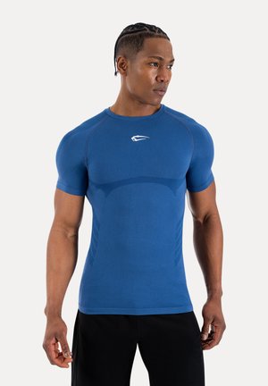 Hombre atlético con cabello trenzado, vestido con una camiseta deportiva ajustada de manga corta azul y pantalones cortos negros, mirando hacia un lado.