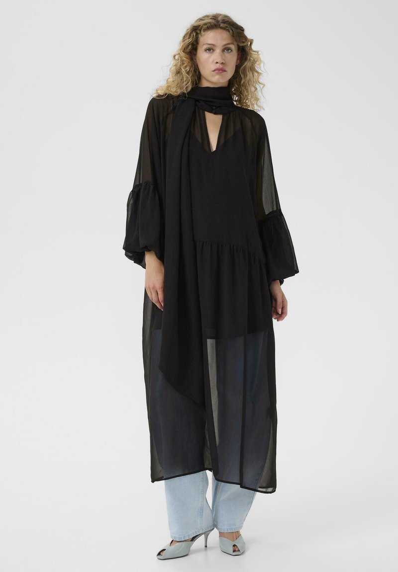 Femme aux cheveux bouclés portant une longue robe noire transparente par-dessus un jean bleu clair et des escarpins ouverts bleu clair, debout devant un fond uni.