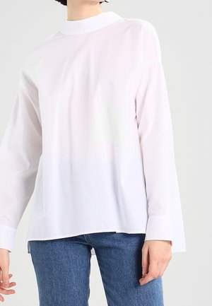 Osoba ubrana w gładką białą bluzkę z długim rękawem i wysokim kołnierzem oraz niebieskie dżinsy, stojąca na tle jasnoszarego tła.