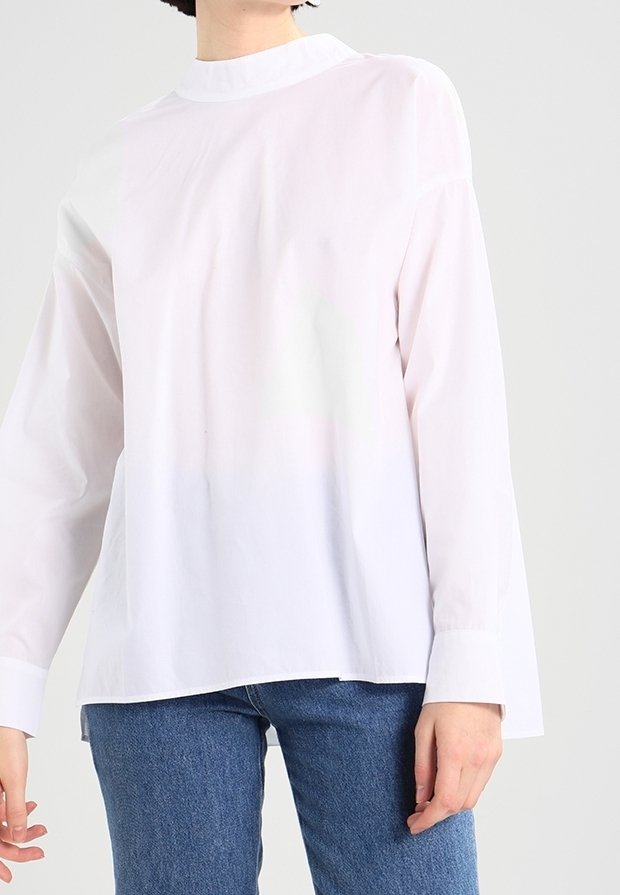 Personne portant un chemisier blanc uni à manches longues avec un col montant et un jean bleu, debout devant un fond gris clair.