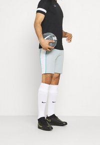 Svart atletisk t-shirt med vita ränder, ljusgrå shorts med teal-accenter, vita strumpor och svarta Nike fotbollsskor, håller en grå boll.