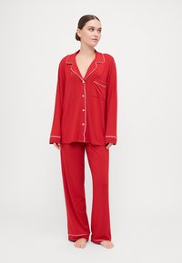 Red pajama set with long sleeves and pants, featuring white piping accents and a chest pocket. Made of soft, smooth fabric.