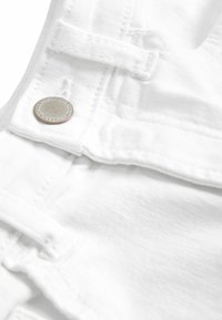 Weiße Denim-Latzhosen verfügen über einen Metallknopf mit der Gravur "AUTHENTIC", verstellbare Träger und eine glatte Stofftextur.