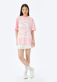 Lyserød oversized t-shirt med hvid grafisk tekst og tropiske designelementer, parret med en hvid plisseret nederdel og hvide sneakers.