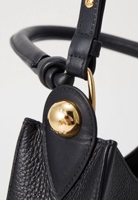 Sac à main en cuir noir avec une finition texturée, doté d'un fermoir circulaire en or et d'une bandoulière nouée. Les détails incluent des coutures apparentes.