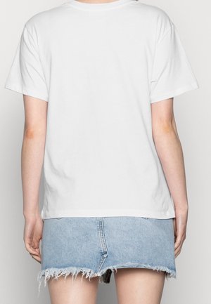 Osoba ubrana w prosty biały t-shirt z krótkim rękawem oraz jasno-niebieską, postrzępioną spódnicę dżinsową, widziana od tyłu.