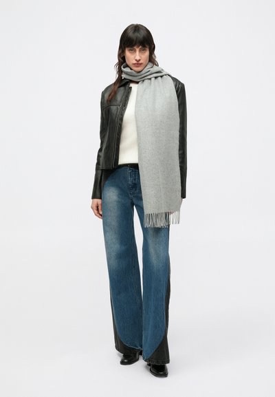 Sciarpa grigia con bordi sfrangiati indossata sopra un maglione bianco e una giacca di pelle nera, abbinata a jeans a gamba larga azzurri chiari e stivali neri.