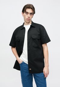 Μαύρο πουκάμισο με κοντά μανίκια, δύο μπροστινές τσέπες και κούμπωμα με κουμπιά, φορεμένο πάνω από λευκό μπλουζάκι, συνδυασμένο με μπλε τζιν τζιν.