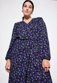 Abito floreale viola con maniche lunghe a sbuffo e scollatura a V. Presenta uno sfondo nero, tessuto testurizzato e dettaglio della vita arricciata.