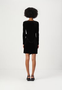 Vestido de veludo preto com mangas longas, corte até ao joelho, decote redondo e textura suave. Usado com sandálias de salto grosso.