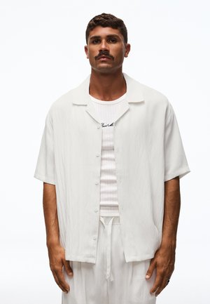 Homme avec moustache porte une chemise blanche ample à manches courtes boutonnée sur un débardeur en maille blanc et un pantalon blanc à cordon, debout devant un fond blanc.
