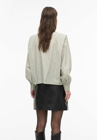 Moteris su ilgais rudos spalvos plaukais, dėvinti kreminės ir juodos spalvų dryžuotą ilgomis rankovėmis palaidinę, juodą odinę sijoną, permatomas pėdkelnes ir juodus batus, žvelgianti į šalį.