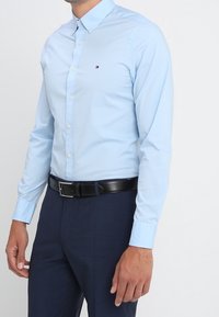 Camisa de botones azul claro, de manga larga y con cuello, que presenta un pequeño logotipo. Combinada con pantalones oscuros y un cinturón de cuero negro.