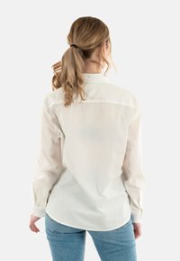 Camicia bianca a maniche lunghe con colletto, caratterizzata da una texture liscia e un orlo curvo, indossata con jeans in denim blu chiaro.