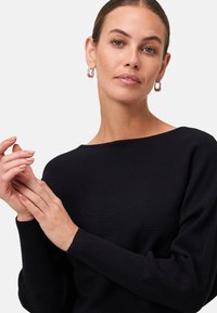 Žena s staženými vlasy má na sobě stříbrné kroužkové náušnice a černý žebrovaný top s dlouhým rukávem, jemně si drží levou ruku pravou.