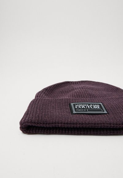 Bonnet en tricot violet avec une texture côtelée et un revers replié. Présente une étiquette rectangulaire noire avec du texte blanc et une bordure décorative.